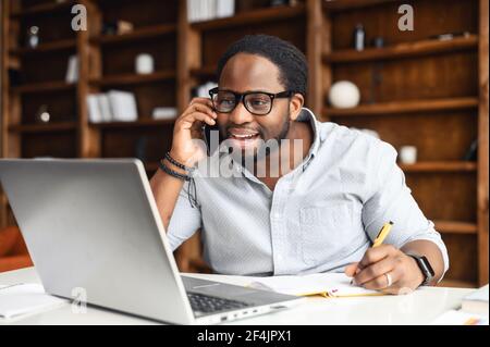 Multitasking African-American businessman office manager in glasses working in the office, sitting at the desk with laptop, holding smartphone, talking with client, looking at the screen, taking notes Stock Photo