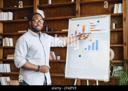 African American male school teacher in glasses pointing on the whiteboard with bar charts, explaining numbers, streaming lesson online and looking at webcam, teaching in a virtual online classroom Stock Photo