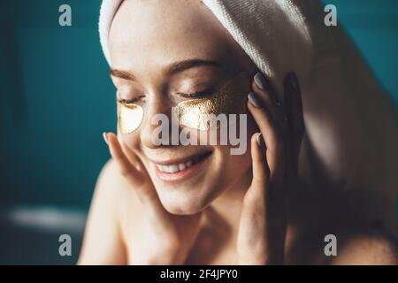 Close up photo of a freckles woman wearing golden eye patches covering her head with a towel and smile Stock Photo