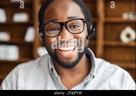 Smiling young African-American salesman in glasses and headset looking at the camera and laughing, positive black man on video call working in the customer service department and talking online Stock Photo