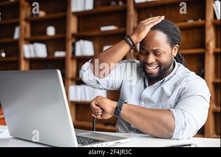 Young African American guy totally forgot about a business meeting, sitting at the desk and working on laptop, checking the time from a watch on his wrist, time to take pills, time for changes concept Stock Photo