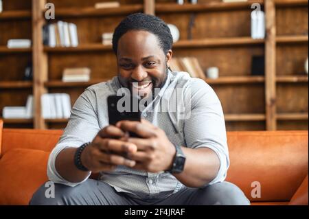 Happy laughing mixed-race man sitting on the couch, staying at home, texting chatting with friends on mobile phone, using dating app, flirting, watching comedy show, funny video, playing games online Stock Photo