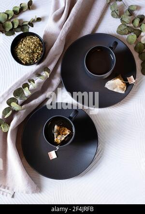 Two identical black tea cups on clay surrounded by eucalyptus branches on a white background Stock Photo