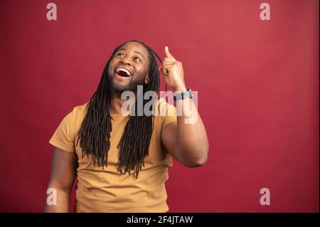 Eureka concept. African American man with dreadlocks standing isolated over red background, pointing with finger in the air, got fresh new business idea, mockup image, open his mouth from excitement Stock Photo