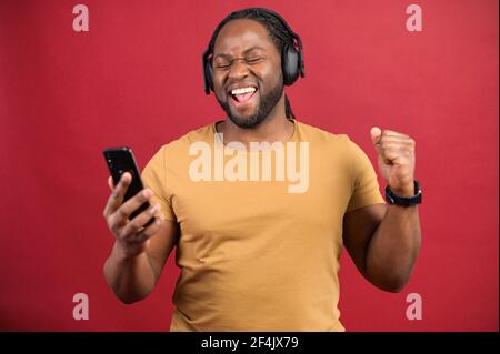 Joyful African-American man wearing wireless headphones holds smartphone in a hand, enjoying music, ecstatic cheerful multiracial guy listening favorite tracks, isolated on red Stock Photo