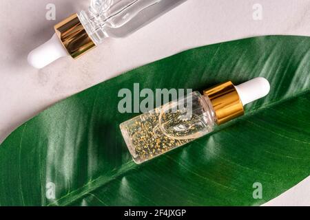 transparent bottle with beauty serum, hyaluronic acid, 24k gold and vitamins on a green tropical leaf. Home face and body care concept Stock Photo