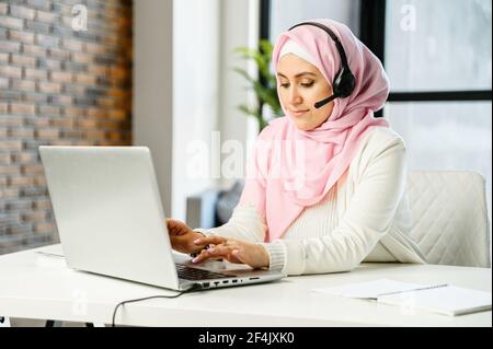 Young Muslim Arab mixed-race female customer service representative in hijab and headset sitting at the desk in office, typing email on laptop, searching or browsing the internet, replying to client Stock Photo