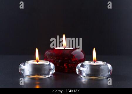 Mourning or prayer concept. Three burning candles on black background. Two of them in transparent glass candlesticks and one in red glass chandelier. Stock Photo