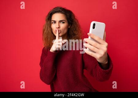Beautiful young woman with curly hair wearing dark red sweater isolated on red background wall holding and using smart phone looking at telephone Stock Photo