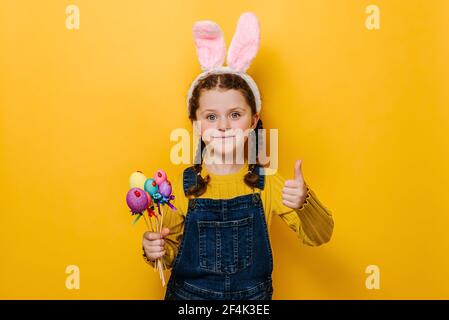 Charming little girl kid holding small colored eggs and showing thumb up as sign of approval or agreement isolated over yellow background Stock Photo