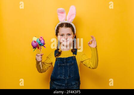 Lovely sweet preschool girl in pink bunny fluffy ears holding colored eggs on sticks and makes okay gesture, agrees with good proposal Stock Photo