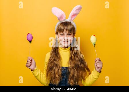 Happy school girl in pink bunny fluffy ears holding two colored eggs, being symbol of religious holiday, isolated on yellow studio background Stock Photo