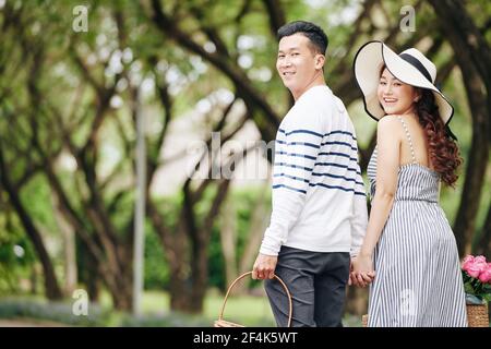 Happy young Asian couple holding hands when walking in park and turning back to camera Stock Photo