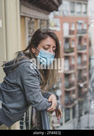 Beautiful depressed lonely woman with face mask in quarantine staring out on balcony feeling sad, lonely, pain and grief during coronavirus outbreak. Stock Photo