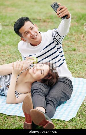Handsome young Vietnamese man taking photo with girlfriend resting head on his laps when they are sitting in park Stock Photo