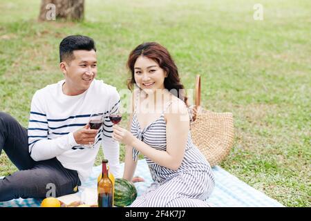 Happy young Asian man with glass of wine looking at his beautiful smiling girlfriend when they are enjoying romantic picnic Stock Photo