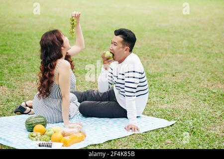 Young Asian couple eating grapes and apples when sitting on blanket in city park Stock Photo