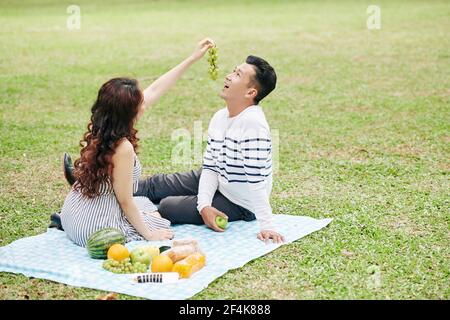 Pretty young woman feeding boyfriend with grapes when they are sitting on blanket in city park Stock Photo