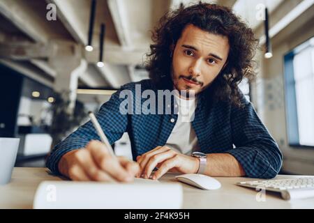 Handsome curly guy is making notes at the working table in open space office Stock Photo