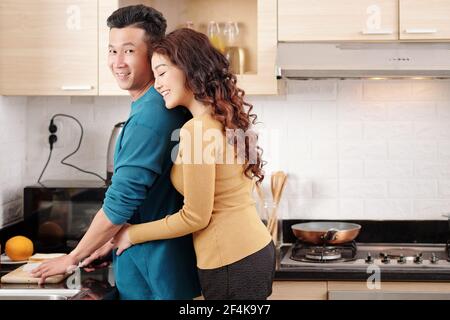 Happy pretty young smiling Vietnamese woman hugging boyfriend cooking breakfast at kitchen counter Stock Photo