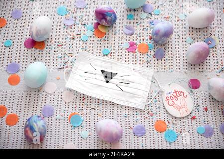 Easter composition with medical mask with painted Easter bunny face and colorful Easter eggs among confetti. Stock Photo