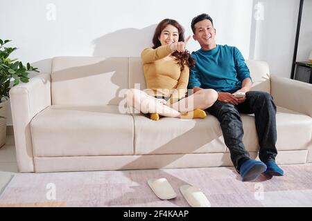 Happy young Asian boyfriend and girlfriend sitting on sofa at home and watching new show on tv Stock Photo