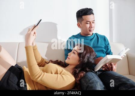 Pretty young Asian woman taking selfie when resting head on laps of boyfriend reading book Stock Photo