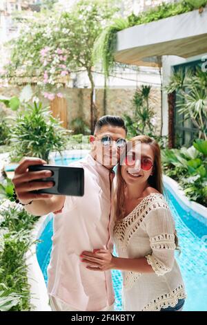 Smiling happy couple in sunglasses standing at swimming pool of spa hotel and photographing on smartphone Stock Photo