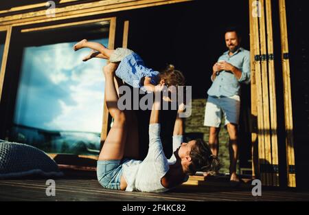Family with small daughter having fun on patio of wooden cabin, holiday in nature concept. Stock Photo