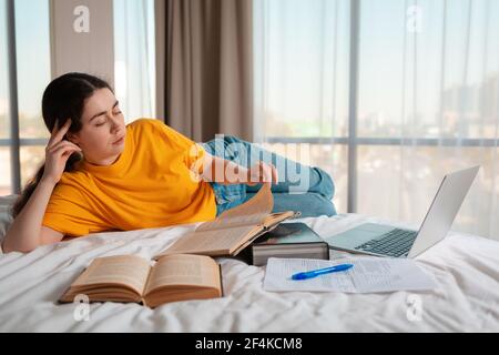 Education. A young woman is lying on her bed reading a book. Laptop and documents are lying next to each other. The concept of online courses and remo Stock Photo