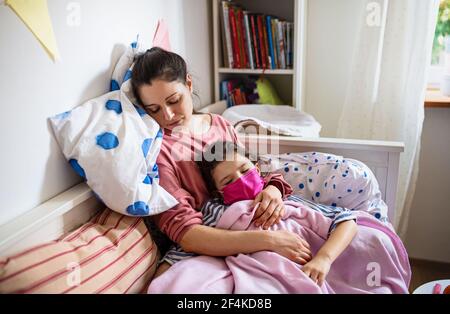 Tired mother looking after sick small daughter in bed at home, coronavirus concept. Stock Photo