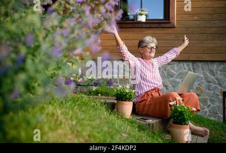 Happy senior woman with laptop working outdoors in garden, home office concept. Stock Photo