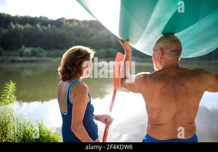 Rear view of senior couple carrying paddleboard by lake in summer. Stock Photo