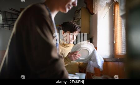 Poor mature mother and small daughter washing dishes indoors at home, poverty concept. Stock Photo
