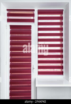 Red or magenta fabric roller blinds on the plastic window and balcony door. Window roller, duo system day and night. Modern shades on apartment window. Empty window sill and light walls. Vertical shot Stock Photo