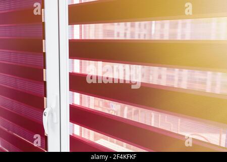 Evening sunlight enters the room through the blinds. Details of red fabric roller blinds on the plastic window in the room or kitchen. Window roller, duo system day and night. Dual roller shades  Stock Photo