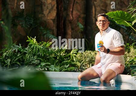 Portrait of handsome fit man in glasses sitting on edge of swimming pool and drinking delicious sweet fruit cocktail Stock Photo