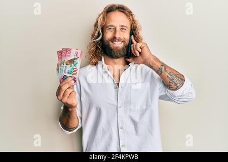 Handsome man with beard and long hair talking on the phone holding 100 new zealand dollars smiling with a happy and cool smile on face. showing teeth. Stock Photo