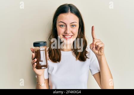 Young brunette woman holding soluble coffee smiling with an idea or question pointing finger with happy face, number one Stock Photo