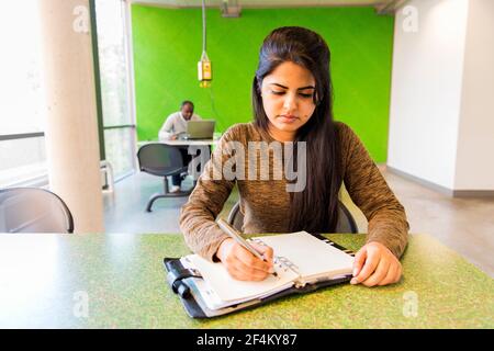 Aachen, Germany. Young, female Indian university exchange student working on a dormatory work desk, using her smartphone and analog notebook. Stock Photo