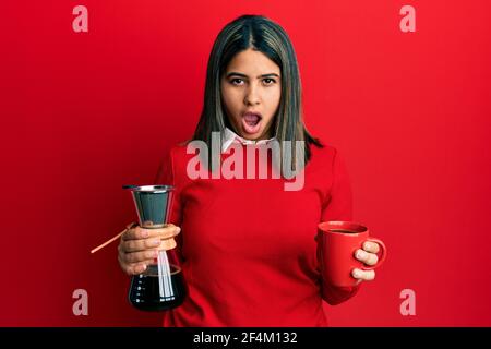 Young latin woman holding coffee filter and cup in shock face, looking skeptical and sarcastic, surprised with open mouth Stock Photo