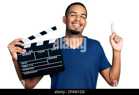 Young african american man holding video film clapboard smiling with an idea or question pointing finger with happy face, number one Stock Photo