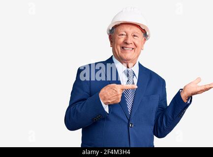 Senior handsome grey-haired man wearing suit and architect hardhat amazed and smiling to the camera while presenting with hand and pointing with finge Stock Photo
