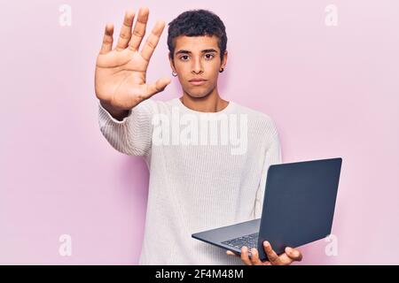 Young african amercian man holding laptop with open hand doing stop sign with serious and confident expression, defense gesture Stock Photo