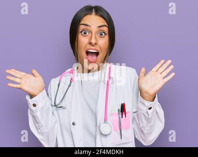 Beautiful hispanic woman wearing doctor uniform and stethoscope celebrating crazy and amazed for success with arms raised and open eyes screaming exci Stock Photo