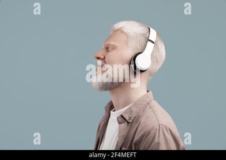 Calm albino guy enjoying music through wireless headphones with closed eyes and smile, listening favorite melodies or audio meditation Stock Photo