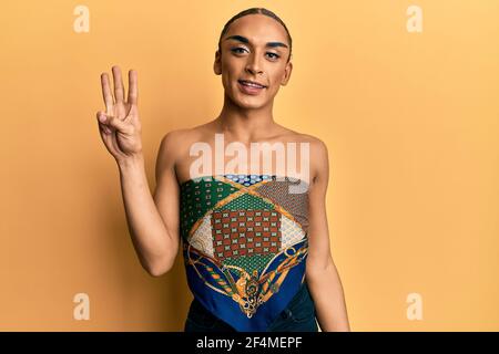 Hispanic man wearing make up and long hair wearing womans top showing and pointing up with fingers number three while smiling confident and happy. Stock Photo
