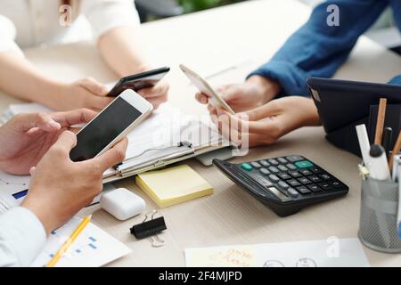 Hands of business people sitting at meeting table with charts and reports and using applications on smartphones Stock Photo