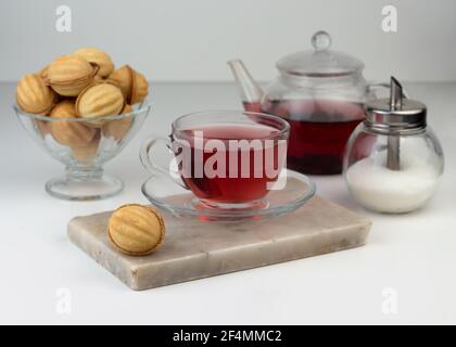 Homemade nut-shaped cookies stuffed with condensed milk in a glass vase on a marble cutting board. Red Sudanese rose tea in a cup and teapot. Stock Photo