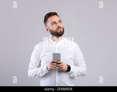 Young businessman with mobile phone deep in thought, looking aside on grey studio background Stock Photo